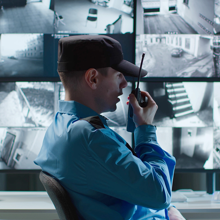 Security guard talking on walkie talkie while looking at cctv camera footage on multiple computer screen. 