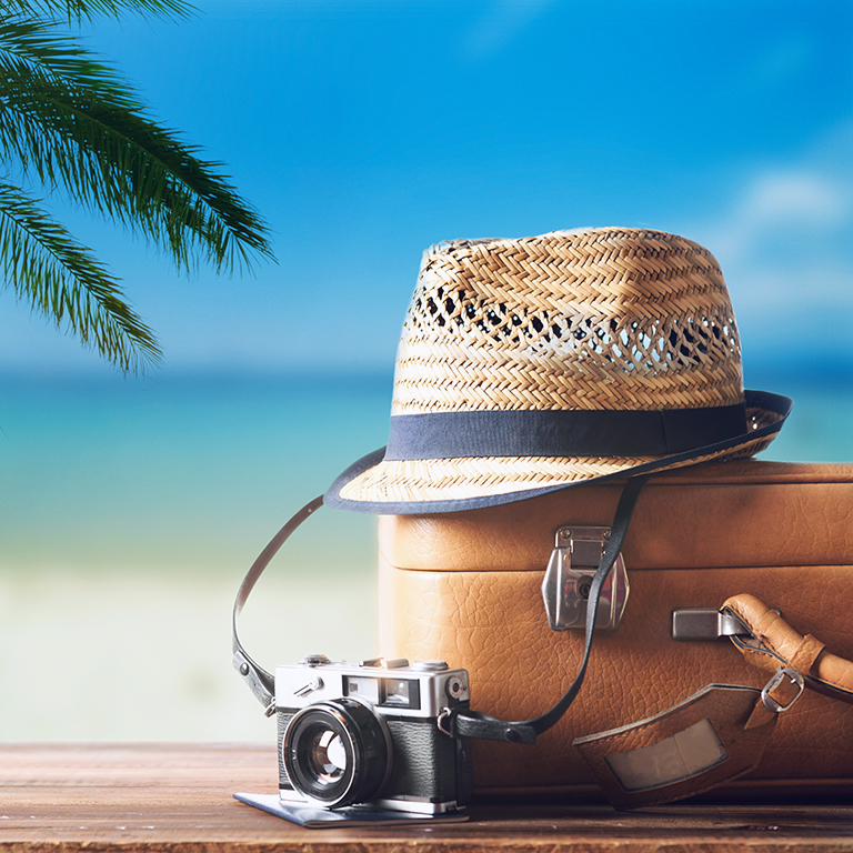 Vintage suitcase, hipster hat, photo camera and passport on wooden dack. Tropical sea, beach and palm three in background. Summer holiday traveling design concept.