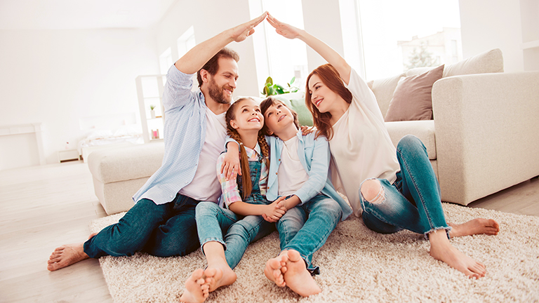 Happy family with two kids sitting on carpet, mom and dad making roof figure with hands arms over heads. New building residential house purchase apartment concept. 